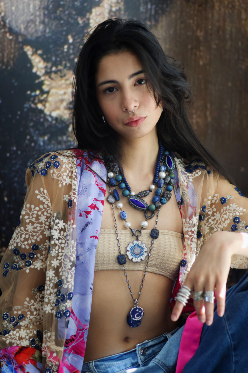 A person wearing a floral shirt and blue jeans, adorned with a stack of blue and gold necklaces featuring lapis lazuli and blue agate.