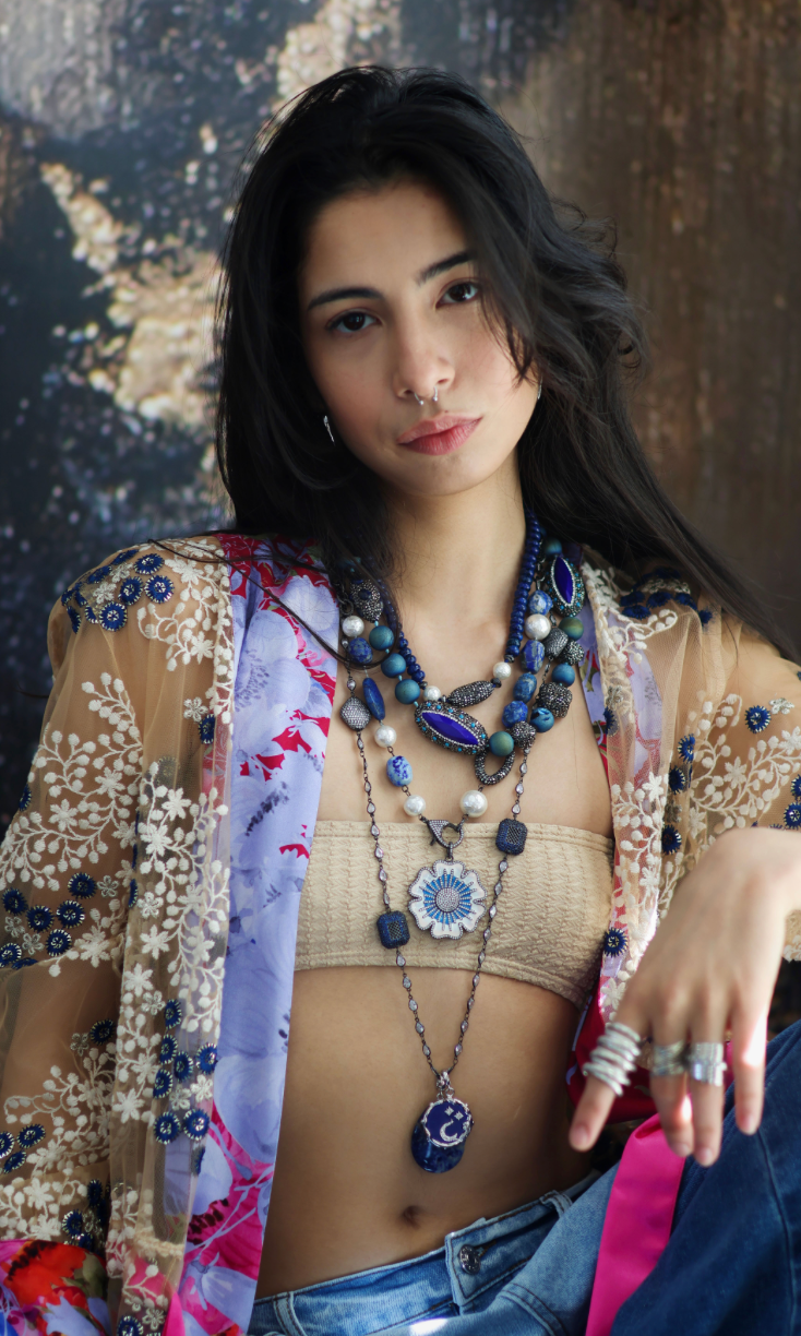 A person wearing a floral shirt and blue jeans, adorned with a stack of blue and gold necklaces featuring lapis lazuli and blue agate.