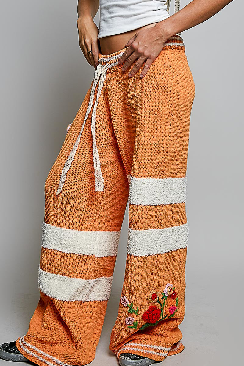Orange and white striped wide-leg knit pants with a drawstring waist and floral embroidery near the hem, shown on a model against a light gray background.
