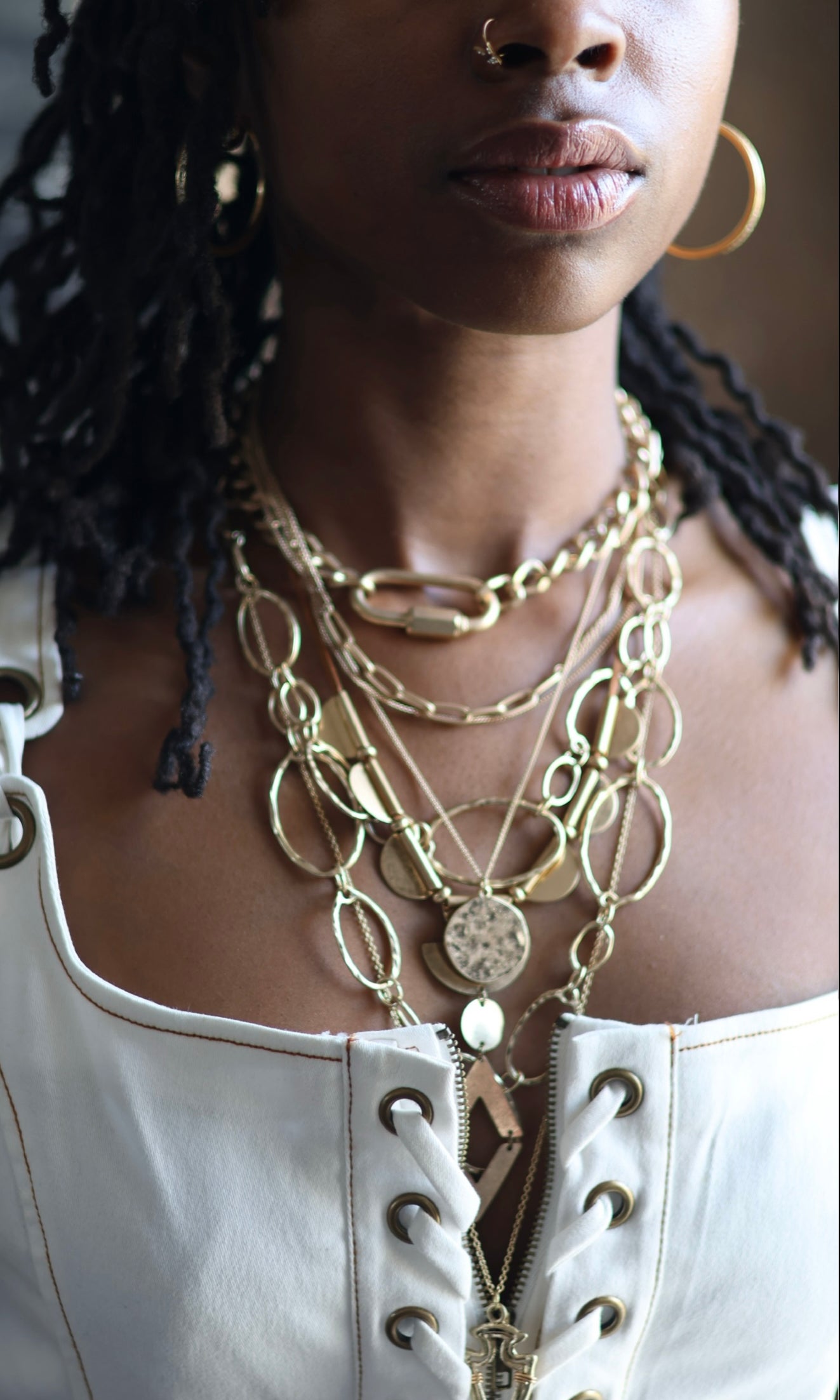 A person wearing a white laced top adorned with multiple gold-colored necklaces featuring various shapes and designs.