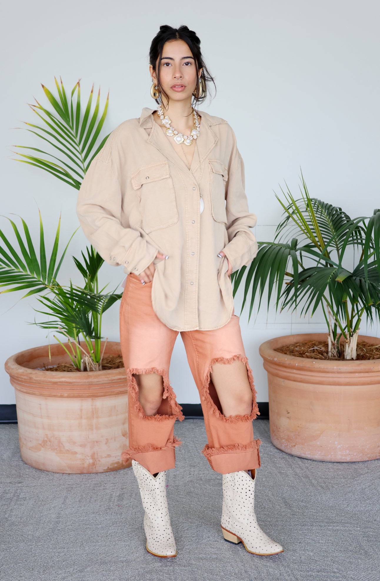 Woman in beige shirt and pink pants standing between two potted plants with a light beige wall background