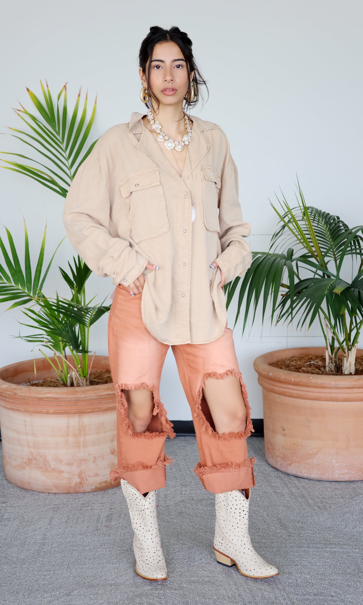Woman in beige shirt and pink pants standing between two potted plants with a light beige wall background