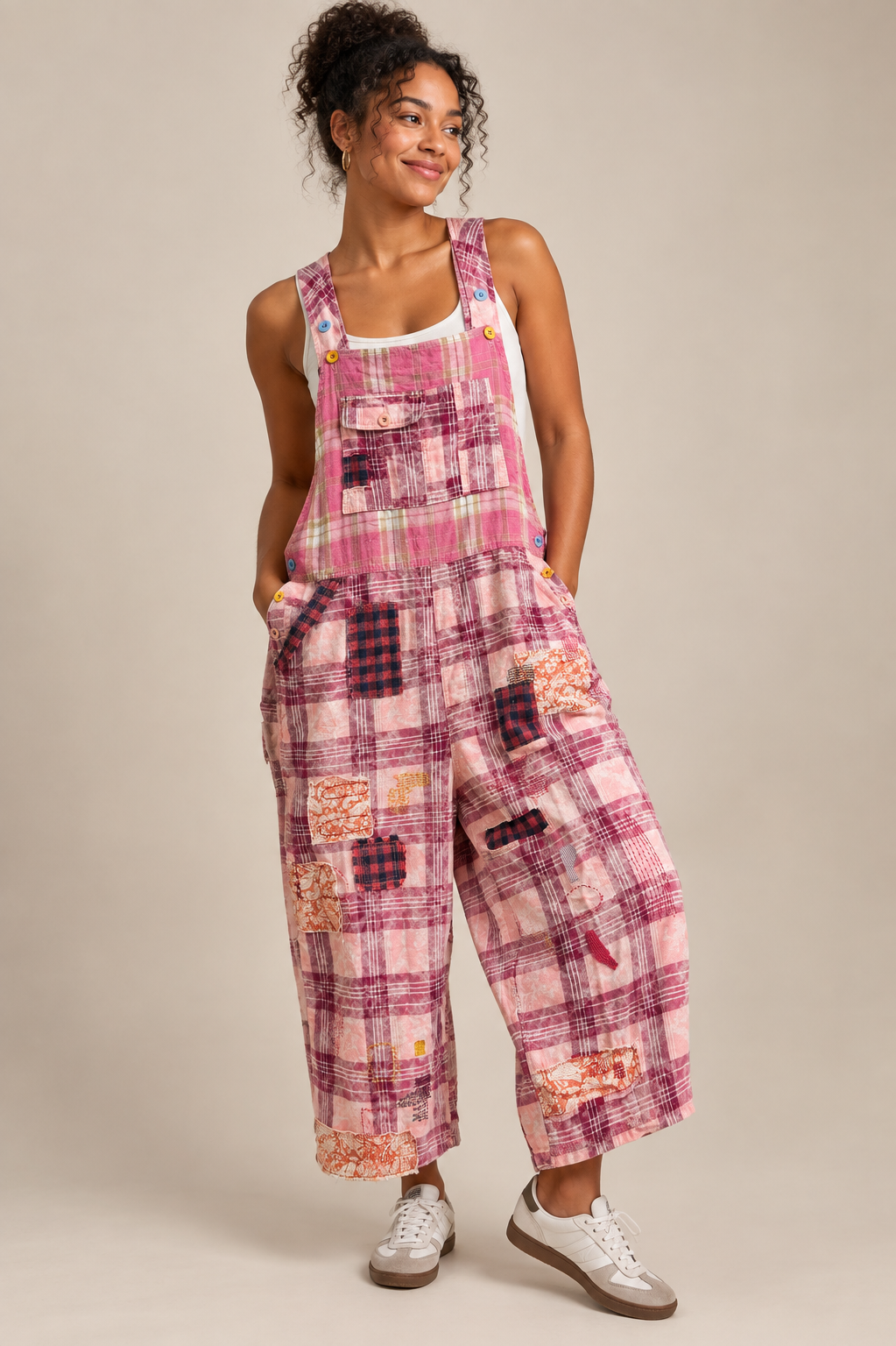 A woman modeling a pink patchwork plaid overall jumpsuit with wide legs, styled with white sneakers against a neutral beige studio background.
