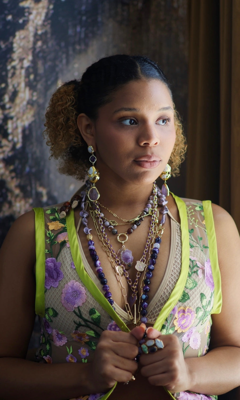 A person wearing multiple necklaces with amethyst stones and gold accents. The necklaces are part of a stack, and the person is holding some of the necklaces, showcasing the jewelry.