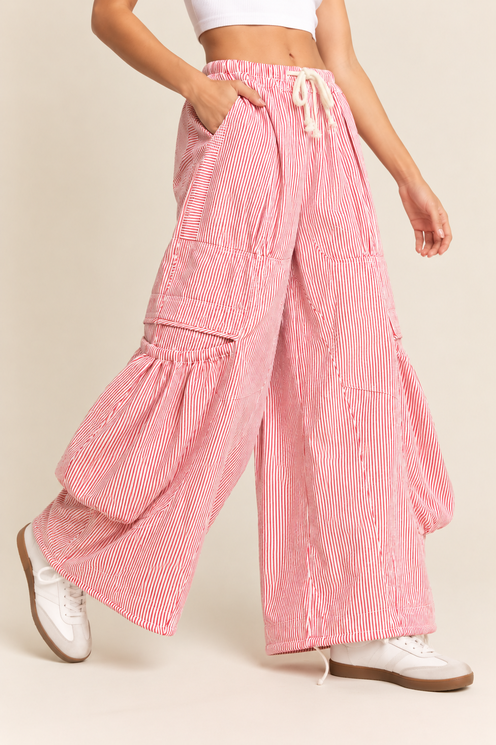 Pink and white pinstriped wide-leg cargo pants with a drawstring waist, oversized utility pockets, gathered panel details, and a relaxed silhouette, styled with a white cropped tank top and sneakers.