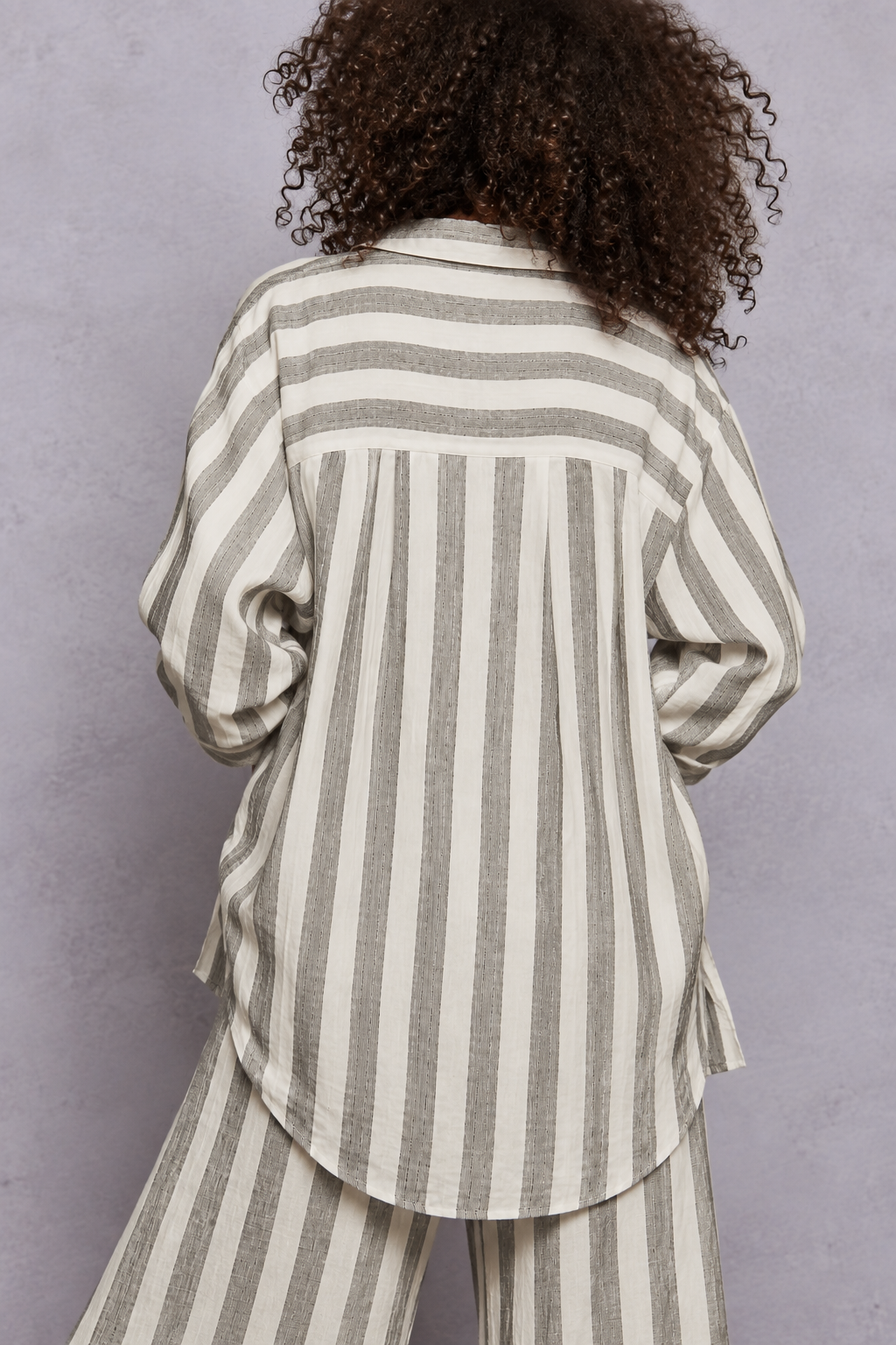 Grey and white vertical striped button-down shirt with rolled sleeves, layered over a white lace-trim camisole and paired with matching striped pants, styled on a model with curly hair.