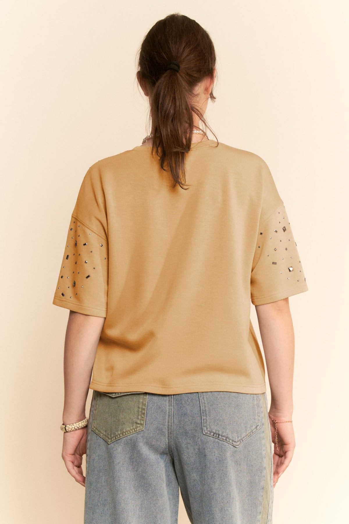 Person wearing a beige t-shirt with embellishments on a plain background