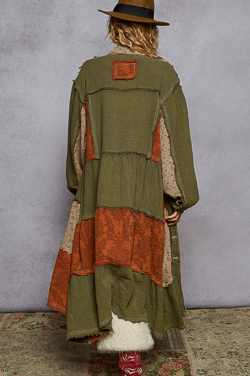 Person wearing a green and orange patchwork coat with a hat, standing against a plain background.