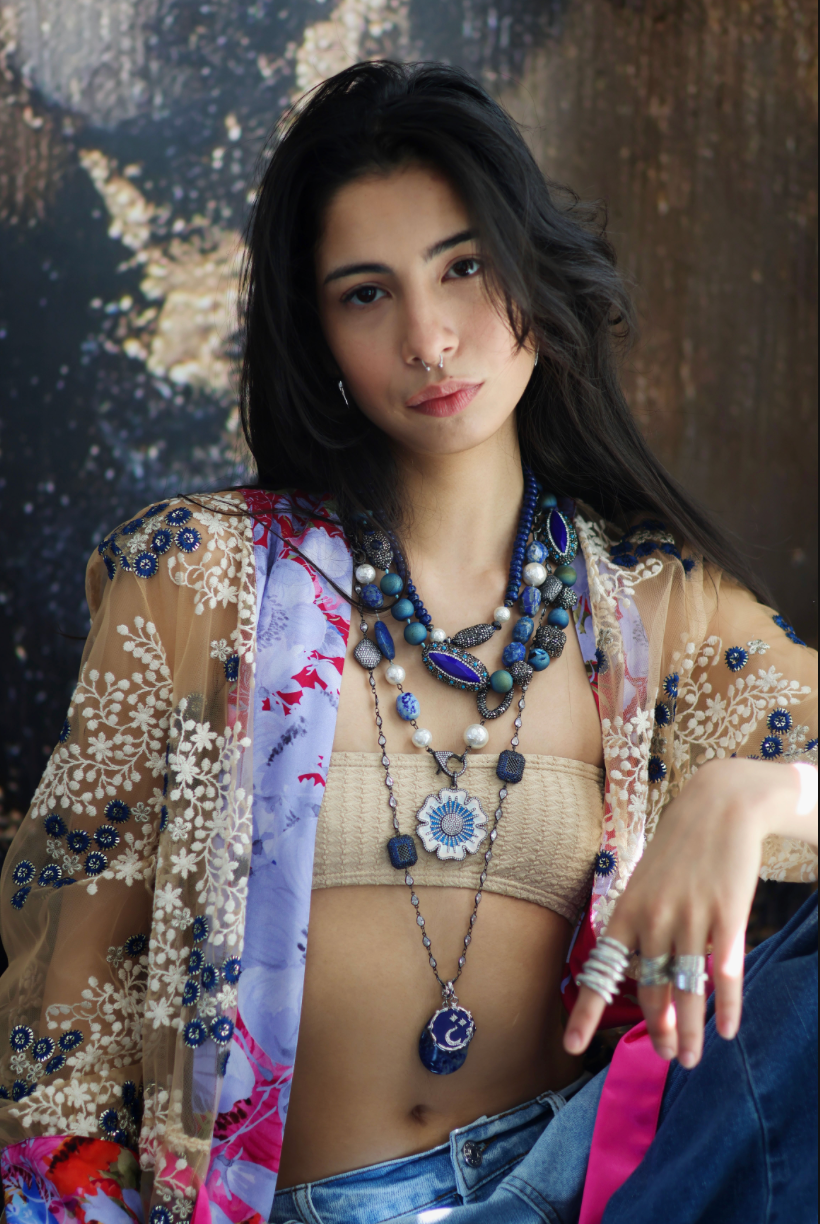 A person wearing a floral shirt and blue jeans, adorned with a stack of blue and gold necklaces featuring lapis lazuli and blue agate.