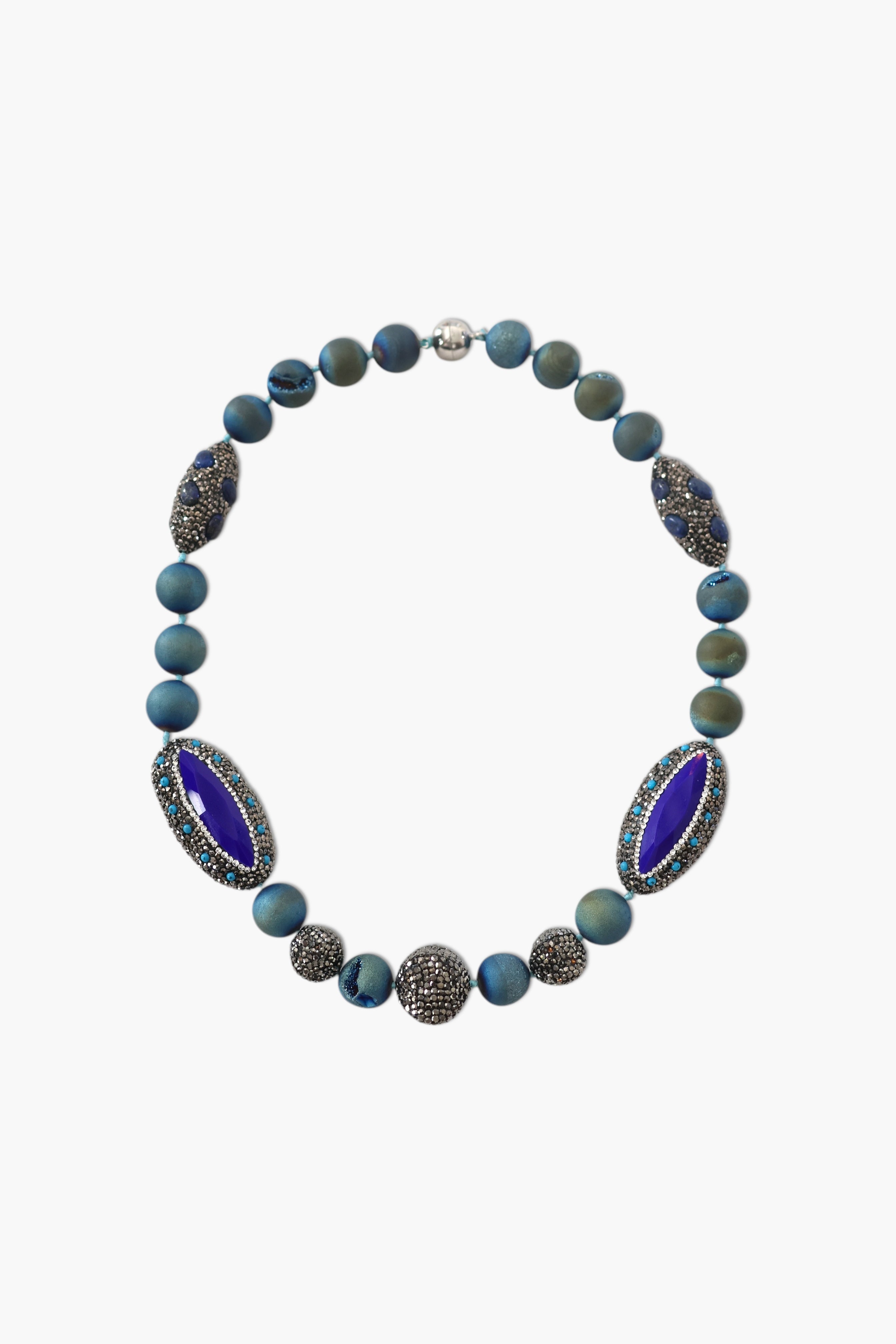 A person wearing a floral shirt and blue jeans, adorned with a stack of blue and gold necklaces featuring lapis lazuli and blue agate.