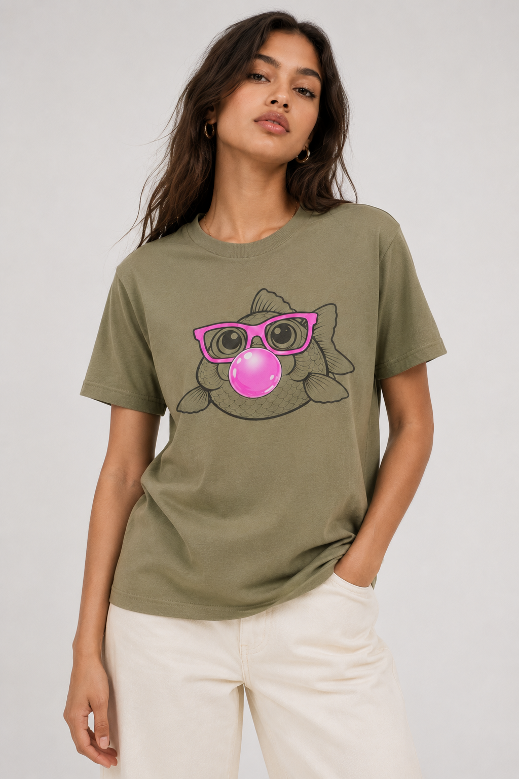 A woman wearing an olive green graphic t-shirt with a cartoon cat wearing pink sunglasses and blowing a pink bubblegum bubble, paired with off-white pants, against a light gray studio background.