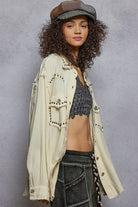 Woman wearing a beige jacket with studded details, gray top, and dark pants against a gray background