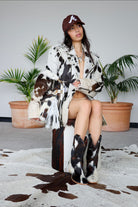 Person wearing a cow print coat and hat, sitting on a stool with plants in the background.