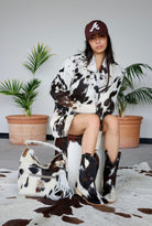 Person wearing a cow print outfit with a matching bag and boots, sitting on a chair.
