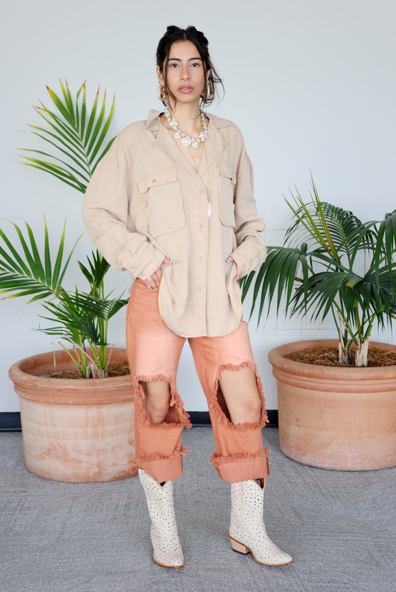 Woman in beige shirt and pink pants standing between two potted plants with a light beige wall background
