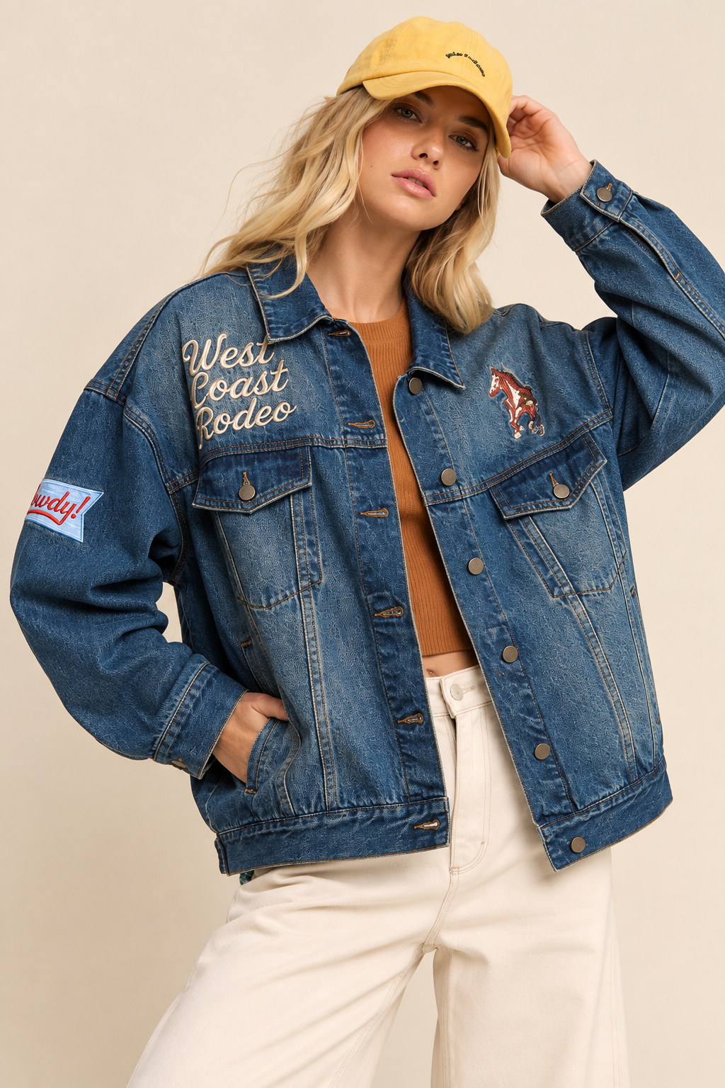 Blue denim jacket with Western-inspired embroidered lettering, horse graphic, and sleeve patches, modeled with a yellow baseball cap, brown top, and cream pants against a neutral studio background.