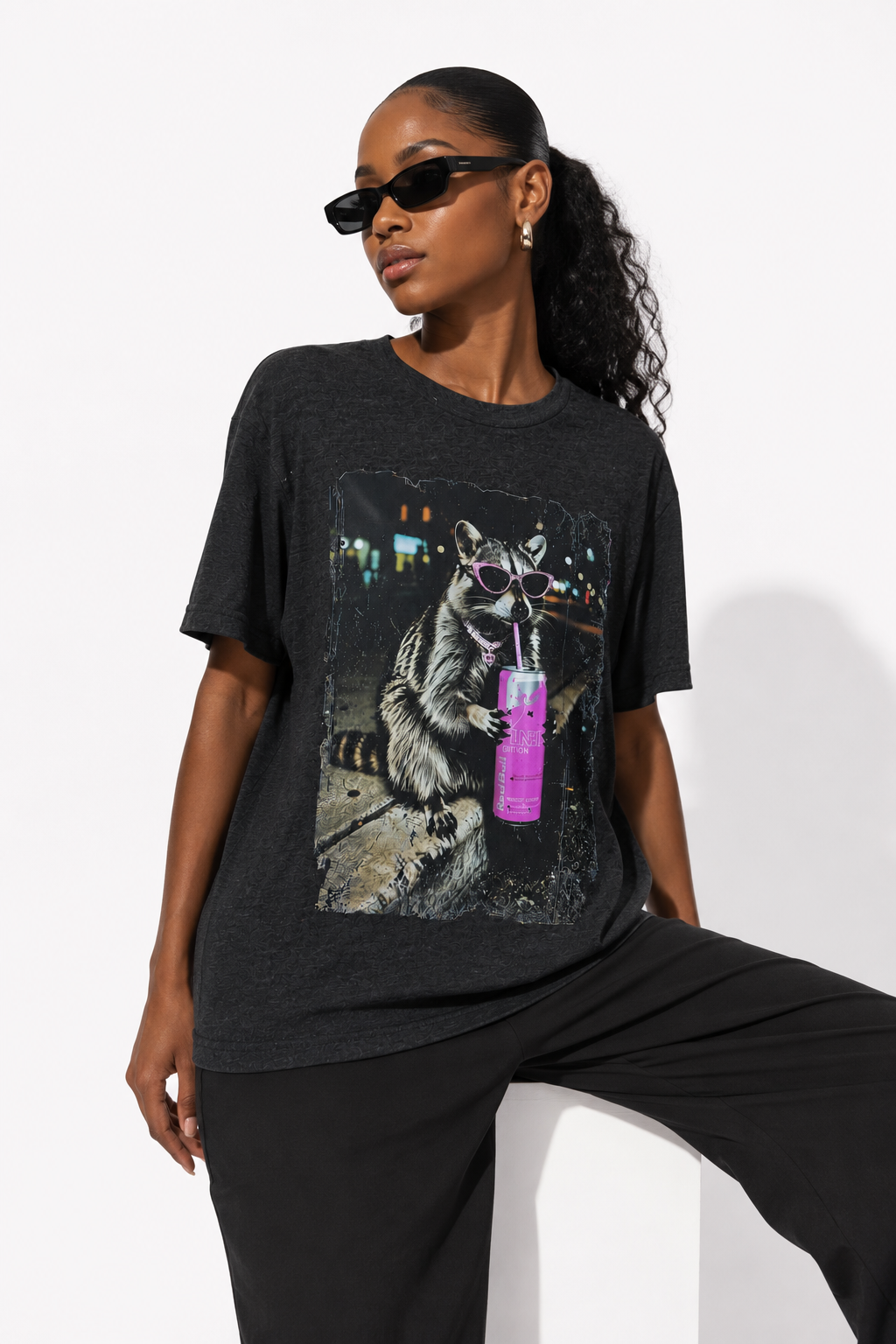 A woman wearing a dark charcoal heather graphic t-shirt with a raccoon holding a pink drink, paired with black pants and sunglasses, posing against a white studio background.