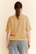 Person wearing a beige t-shirt with embellishments on a plain background