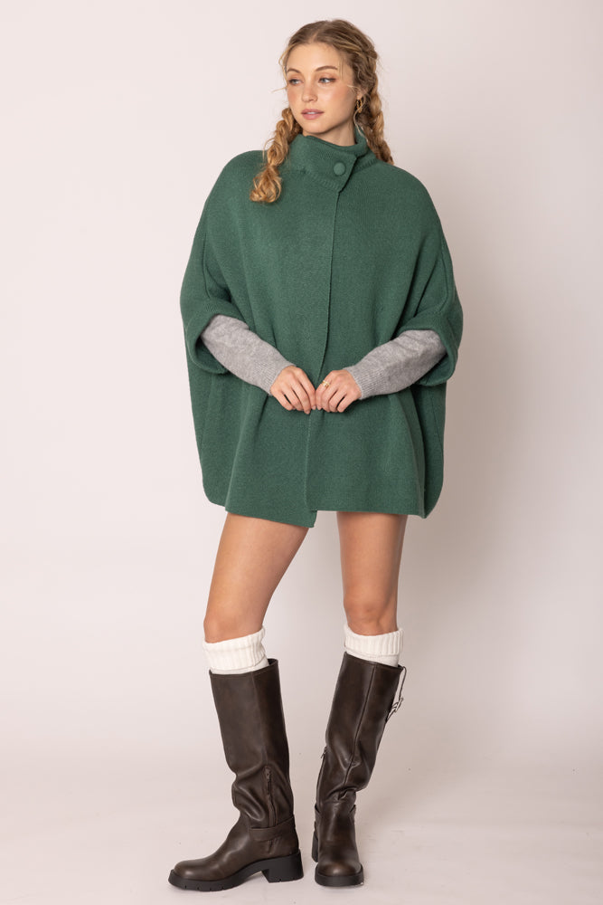 Woman wearing a green poncho with gray cuffs and sleeves, paired with brown boots on a white background.