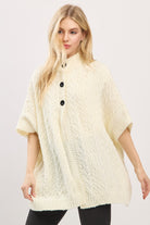 Woman wearing a cream-colored poncho with black buttons on a plain background